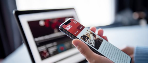 Foto Mensch mit Mobiltelefon in der Hand vor dem Computer